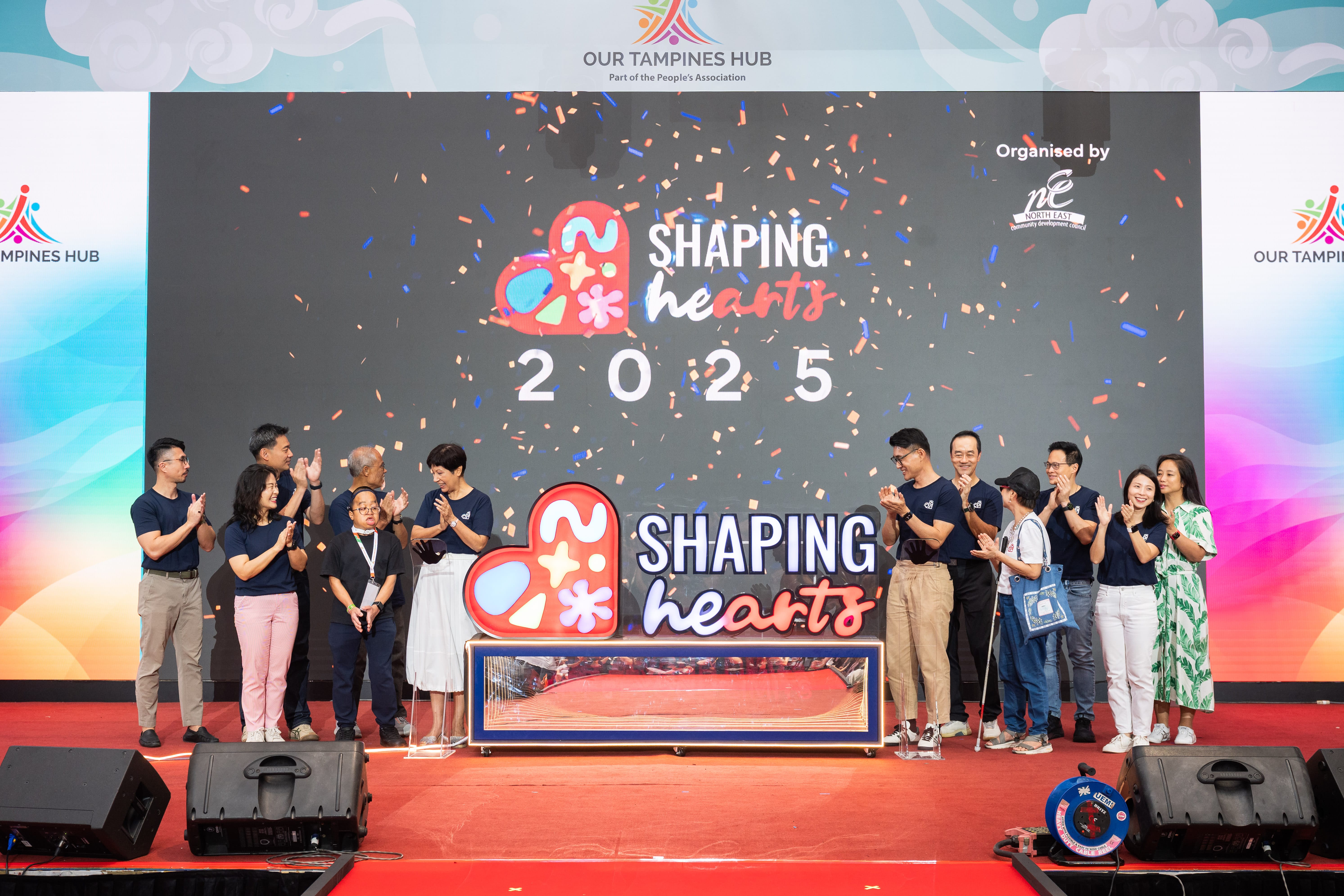 People standing on either side of a ‘Shaping Hearts 2025’ lightbox, clapping and smiling as confetti falls, celebrating the launch of the event.